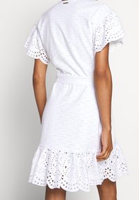 Abito bianco in tessuto con trafori, maniche corte e orlo arricciato. Presenta una vita con cintura e motivi a intaglio in tutto l'abito.