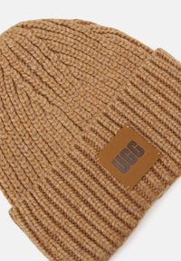 Brun strikket lue med ribbestrikket design, laget av mykt materiale, med en rektangulær lærpatch som viser "UGG." Strukturert overflatedetalj.