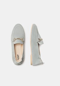 Mocassini in suede blu chiaro con una texture morbida, dettagli in metallo dorato e suola piatta beige. Progettati per comfort e facilità d'uso.