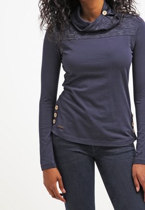 Kobieta ubrana w granatowy top z długim rękawem i kołnierzem typu komin, z drewnianymi guzikami na ramionach i bokach, zestawiony z ciemnymi dżinsami.