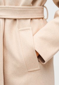 Κοντινή λήψη ενός μπεζ μάλλινου παλτού με ζώνη δεμένη στη μέση και ένα χέρι που είναι εισαγμένο σε ένα ορθογώνιο πλαϊνό τσέπη.