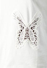 Bílá bavlněná košile s jemným bílým krajkovým designem motýla uprostřed, která vystihuje složité vzory a detailní textura.