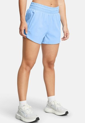 Athlet trägt hellblaue Sportshorts, weiße Knöchelsocken und weiße Laufschuhe und steht vor einem schlichten Hintergrund.