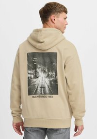 Beige kapucnis pulóver, nagy fekete-fehér grafikával a hátsó részén, amely egy városi utcát ábrázol. Az alján a "BLEND SINCE 1993" felirat található.