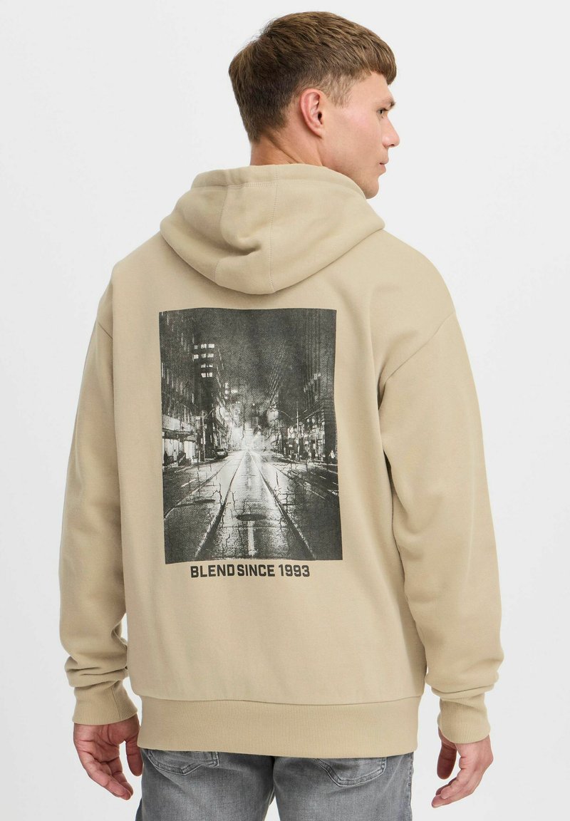 Beige kapucnis pulóver, nagy fekete-fehér grafikával a hátsó részén, amely egy városi utcát ábrázol. Az alján a "BLEND SINCE 1993" felirat található.