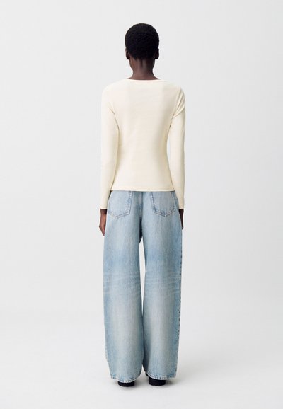 Persona con capelli corti che indossa un top a maniche lunghe color crema e jeans a gamba larga blu chiaro, in piedi voltata verso l'esterno su uno sfondo chiaro.