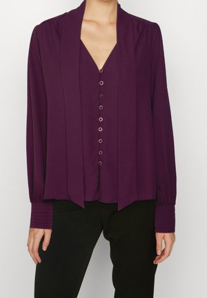 Femme portant un chemisier manches longues violet profond avec un col en V et des accents décoratifs en anneau à l'avant, associé à un pantalon noir.