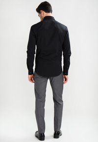 Camicia nera a maniche lunghe con colletto standard, tessuto morbido e polsini con bottoni; abbinata a pantaloni grigi su misura e scarpe nere.
