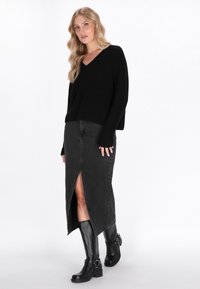 Schwarzer, gerippter Strickpullover mit V-Ausschnitt, kombiniert mit einem schwarzen Jeansrock mit hohem Schlitz und kniehohen schwarzen Stiefeln mit Hardware-Details.