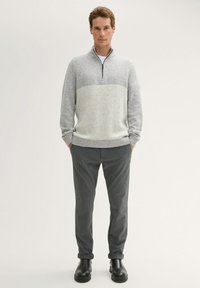Grijze gebreide trui met een rits, voorzien van een lichtgrijze streep over de borst; gecombineerd met donkergrijze broek en zwarte laarzen.