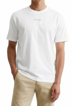 Homme portant un t-shirt blanc uni à manches courtes avec un petit logo « Marc O'Polo » sur la poitrine, accompagné d’un pantalon beige, mains détendues le long du corps.