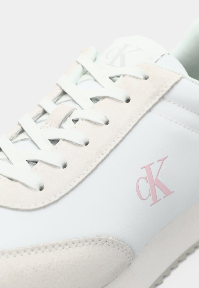 Baskets blanches avec logo rose clair, dotées d'un panneau en cuir suédé texturé, de lacets blancs plats et d'un design minimaliste. Semelle en caoutchouc lisse.