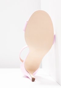 Sandale à talon haut rose clair vue de la semelle, posée contre un mur blanc en angle.