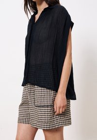 Femme portant un haut noir ample et transparent ainsi que des shorts à carreaux beige et noir, debout devant un fond blanc uni.