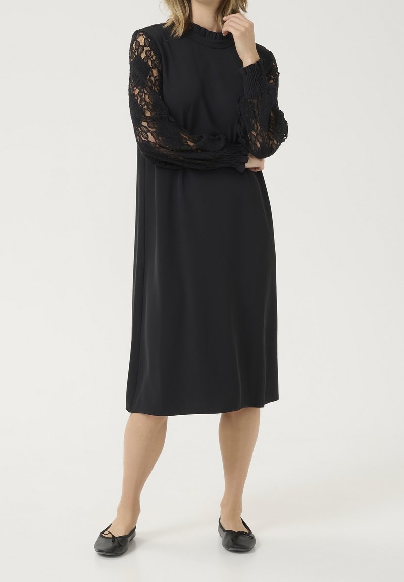 Femme portant une robe noire longueur genou avec des manches longues en dentelle et des ballerines noires, debout devant un fond uni.