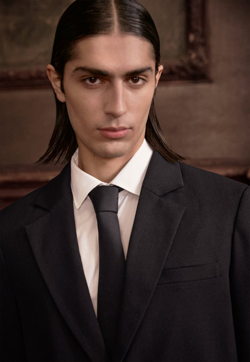 Black suit jacket with a notched lapel, white dress shirt, and black tie; smooth texture, tailored fit, clean lines, and subtle details.