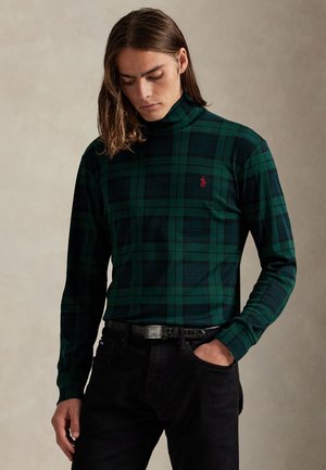 Homme aux cheveux mi-longs, portant un col roulé à carreaux verts et noirs et un pantalon noir, une main dans la poche, regardant vers le bas.