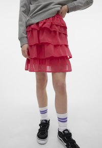 Röd lagerkjol i genomskinligt tyg med volanger. Buren med svarta sneakers och vita strumpor med lila ränder.