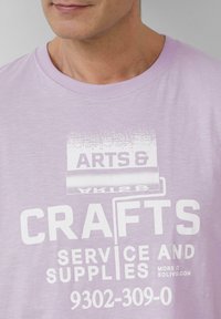 Licht paarse katoenen t-shirt met witte tekst die leest "KUNST EN HANDWERK SERVICE EN BENODIGDHEDEN" met een roller graphic.