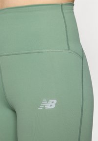 Zelené legíny s vysokým pásom vyrobené z elastického materiálu, s kontrastným prešívaním a reflexným logom New Balance na spodnej strane.