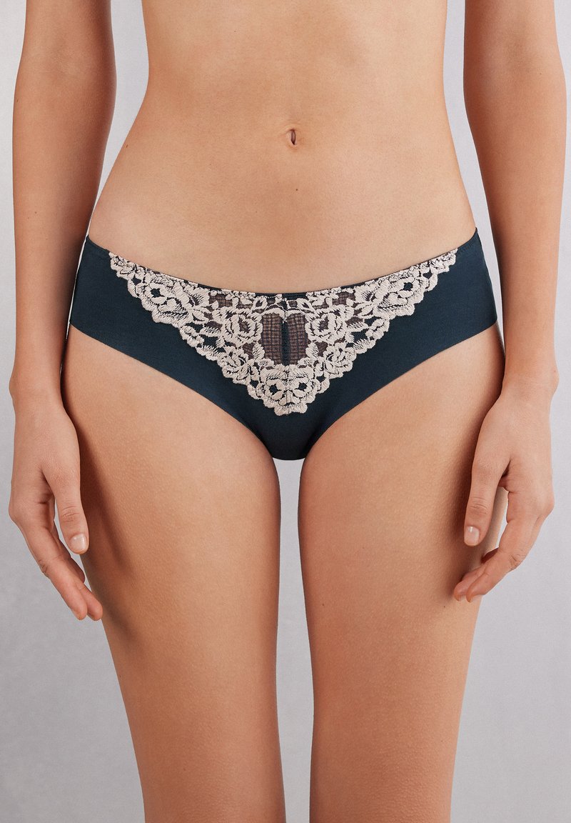 Ciemnozielona bielizna z koronkowymi wykończeniami, ozdobiona delikatnym beżowym haftem kwiatowym z przodu, o gładkiej strukturze materiału i klasycznym kroju majtek.