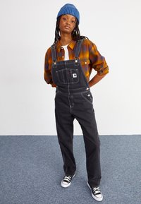 Svarte jeanssalopetter med en frontlomme, sølvfarget hardware og justerbare stropper, kombinert med en oransje og blå rutete skjorte og en blå lue.