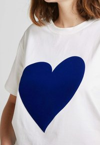T-shirt en coton blanc avec un grand motif de cœur en feutre bleu marine sur le devant. Encolure simple et manches courtes.