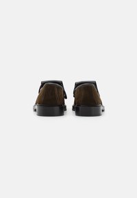 Zapatos de ante marrón con acentos negros, punta redonda, suela plana y un tacón pronunciado. El diseño presenta un estilo minimalista y líneas limpias.