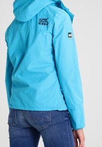 Lichtblauwe waterdichte jas met capuchon, met een logo op de achterkant en mouw. Gecombineerd met donkerblauwe spijkerbroek, met een gladde textuur.