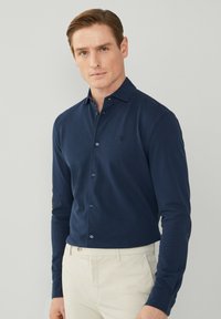 Camicia a maniche lunghe blu navy con colletto a punta, abbottonatura frontale e logo discreto sul petto. Indossata con pantaloni chiari.