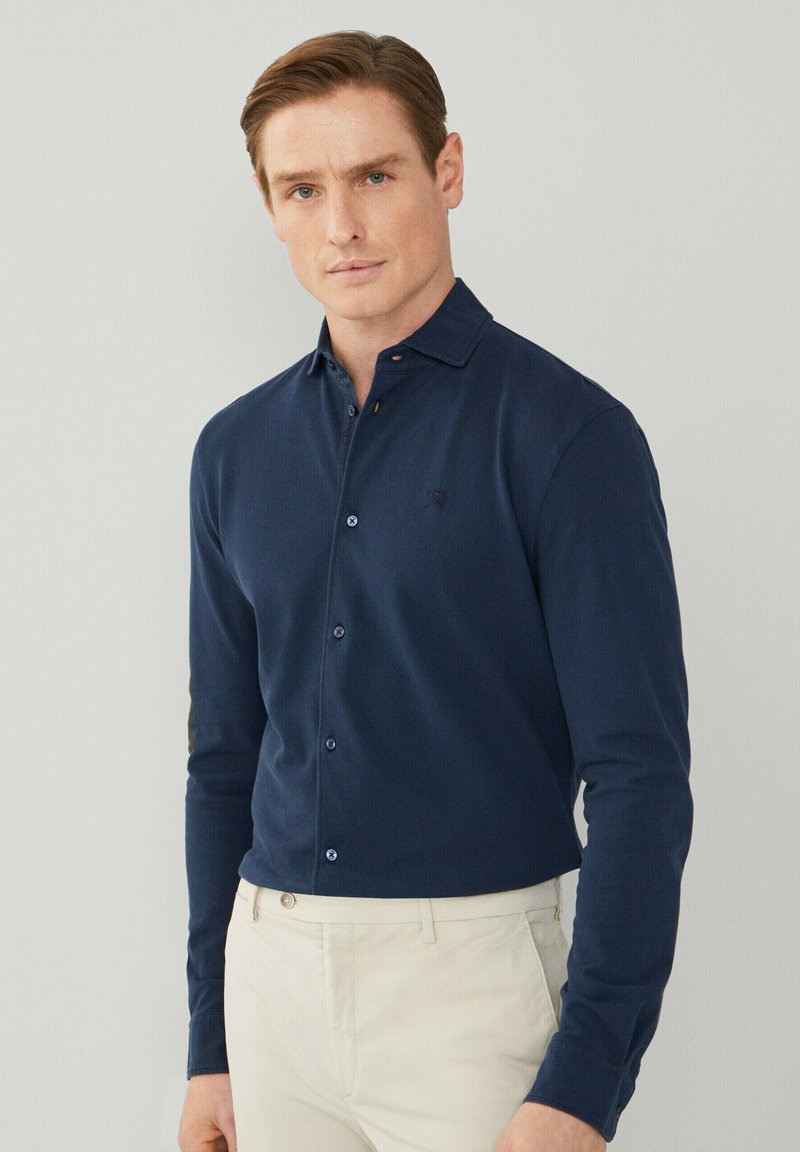 Camicia a maniche lunghe blu navy con colletto a punta, abbottonatura frontale e logo discreto sul petto. Indossata con pantaloni chiari.