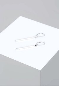 Paire de boucles d’oreilles argentées en forme de goutte avec de fines barres rectangulaires suspendues à des fermoirs à levier sur une surface blanche inclinée.