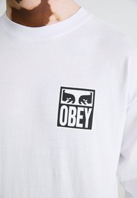 White cotton t-shirt featuring a black square logo with the word "OBEY" and abstract designs above it. Short sleeves and crew neck.