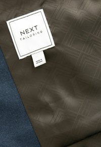 Maßgeschneiderter Jackenstoff in Dunkelgrün mit einer feinen geometrischen Textur, kombiniert mit blauen Stoff. Verfügt über ein weißes Etikett für NEXT Maßanfertigung.