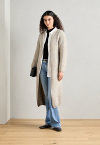Long, light brown cardigan made of soft, textured material, open front design, worn over a black top and paired with light blue jeans.