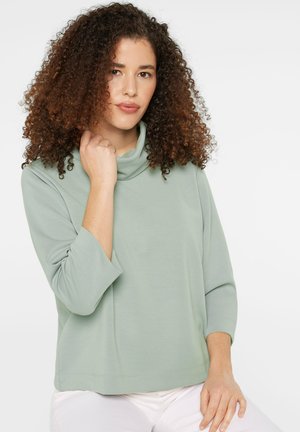 Frau mit lockigem Haar, die ein hellgrünes Langarmshirt und weiße Hose trägt, und sanft den Kragen berührt, sitzt vor weißem Hintergrund.