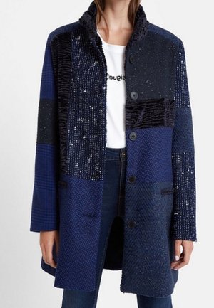 Kobieta w niebieskim płaszczu typu patchwork z różnymi teksturowanymi kwadratami materiału, czarnymi guzikami, na białej koszulce z nadrukiem i ciemnych dżinsach.