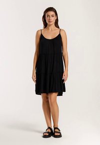 Zwarte gelaagde jurk met dunne bandjes, gemaakt van een lichtgewicht stof. Heeft een losse pasvorm en valt boven de knie. Gecombineerd met zwarte sandalen.