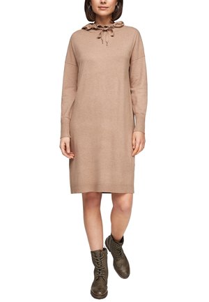 Kobieta w beżowej dzianinowej sukience z długim rękawem, z falbankowym dekoltem i ściągaczem, zestawiona z ciemnooliwkowymi, sznurowanymi botkami za kostkę.