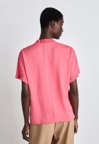 Kurzärmliges, korallenfarbiges Hemd mit einem gefalteten Kragen. Leichter Stoff, leicht lockere Passform und abgerundeter Saum. Rückansicht gezeigt.