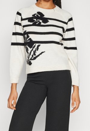Femme portant un pull blanc à rayures horizontales noires et motifs floraux, associée à un pantalon noir, debout devant un fond uni.
