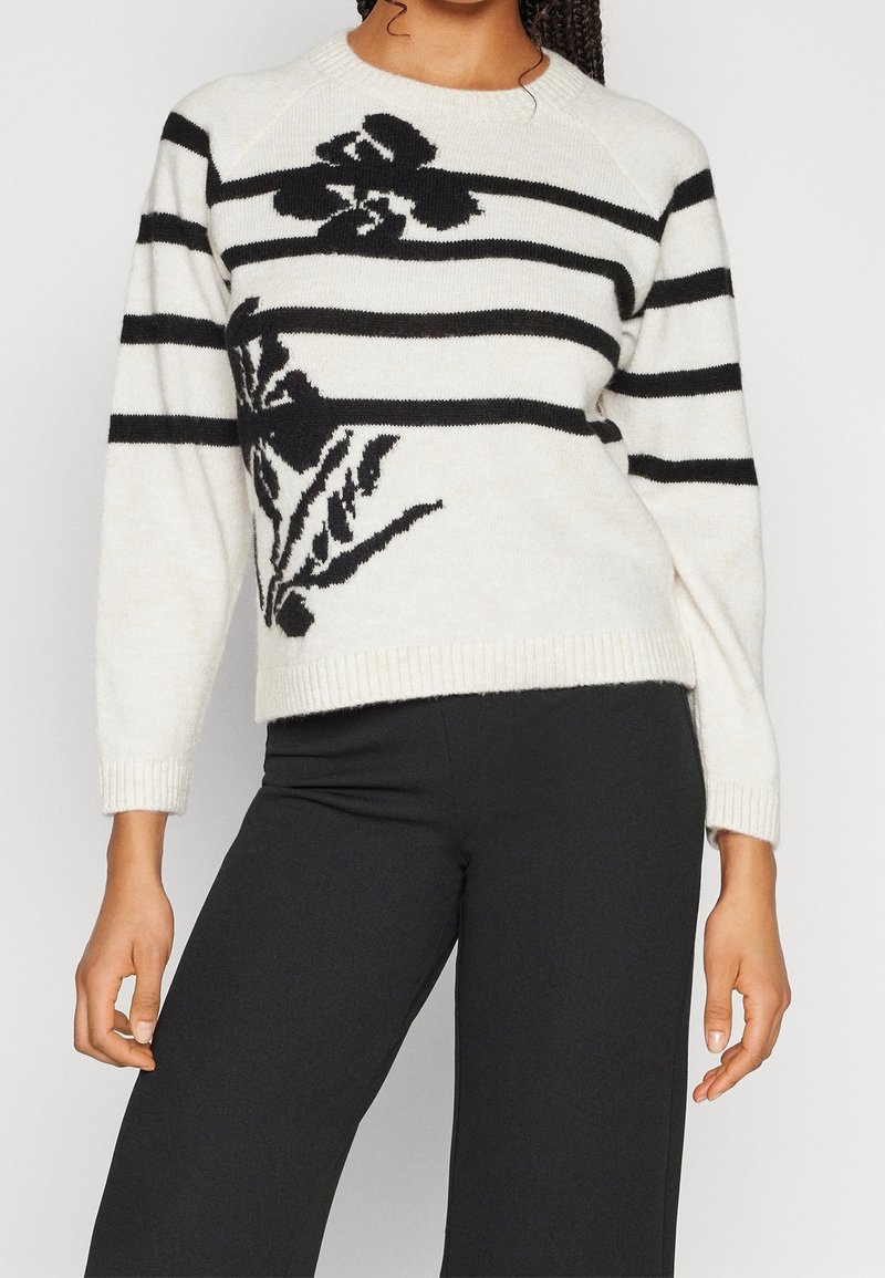 Femme portant un pull blanc à rayures horizontales noires et motifs floraux, associée à un pantalon noir, debout devant un fond uni.
