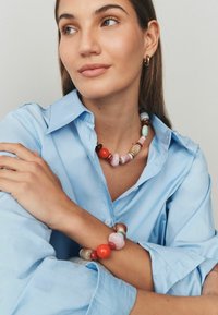 Krāsains pērlīšu kaklarota ar dažādām formām, kurā ir oranžas, rozā un zilas krāsas. Valkāta ar gaiši zilu krekliņu ar pogām.