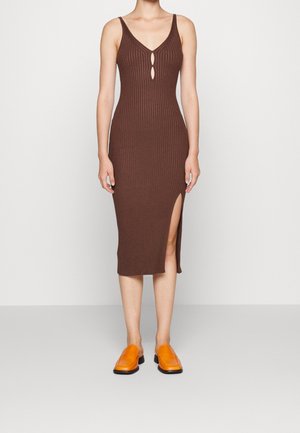 Ærmeløs brun ribstrikket midi-kjole med udskæringer foran og sidelomme, kombineret med lyse orange slip-on sko, stående mod en ensfarvet baggrund.