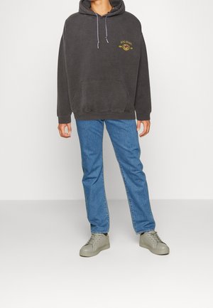 Hoodie gris foncé avec une poche avant, arborant un logo brodé en or. Associé à un jean bleu et des baskets beiges pour un look décontracté.