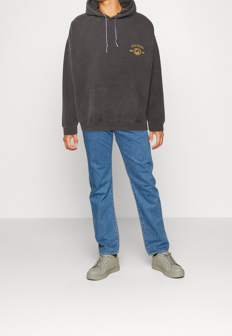 Felpa grigia scura con tasca frontale, con logo ricamato oro. Abbinata a jeans blu e sneakers beige per un look casual.