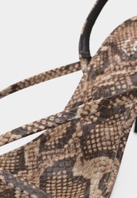 Gros plan sur une sandale à motif peau de serpent marron avec des fines lanières et un design à bout carré sur fond blanc.