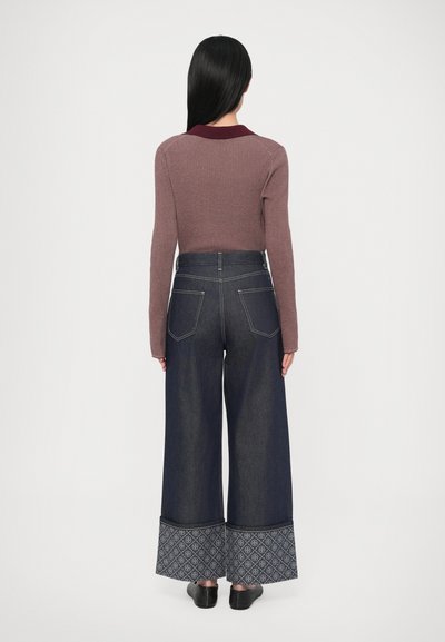 Sarkanīgi brūns ribots garroku tops, kopā ar tumšu džinsa platajiem biksēm ar rakstainiem manžetēm un augstu vidukli, pabeigts ar melnām kurpēm.