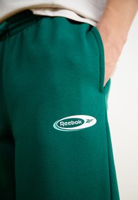 Zelené flísové tepláky s nastaviteľným pásom, s bielym logom Reebok na stehne a bočnými vreckami. Hladká textúra, ležérny dizajn.