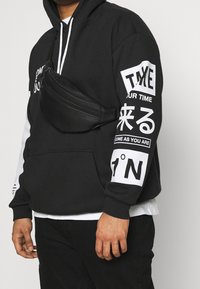 Sweat à capuche noir avec texte graphique blanc et accents sur les manches ; présente un sac banane noir porté sur la poitrine. Matériau doux, coupe décontractée.